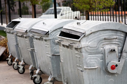 A row of four large metal wheelie bins positioned on a paved surface, likely for rubbish disposal in a residential or commercial area in Staines. The bins are constructed from galvanized steel with a textured, metallic finish and are equipped with black plastic lids, some of which are slightly open. Each bin has four small rubber wheels for mobility, and they are aligned side by side, with the first bin on the left showing a small label or opening mechanism. Behind the bins, a black metal fence separates the area from a street with parked cars, trees, and a partly cloudy sky visible in the background. The overall scene is well-lit with natural daylight, illustrating a typical outdoor waste collection setup. The arrangement and condition of the bins suggest they are used for general waste or rubbish collection services, such as those provided by Garden Clearance Staines, which may operate in the local area around Staines or nearby postcode regions.