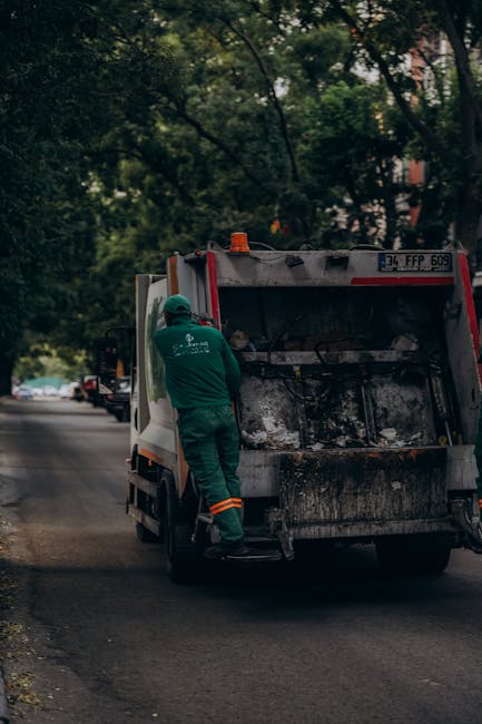 A middle-aged man with short dark hair, wearing a dark T-shirt with white text and dark pants, is seen bending over a modern stainless steel rubbish bin with a rounded top and an open slot for disposa