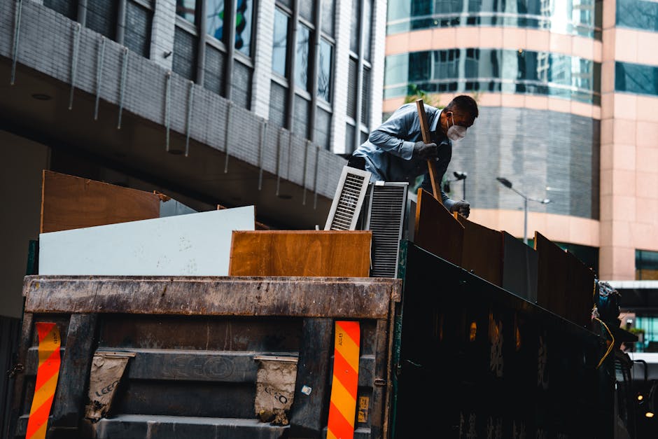 A man wearing a light blue shirt, dark pants, and a face mask is standing on the edge of a large, open-top rubbish truck, loading or sorting various discarded items. The truck contains a mix of materi