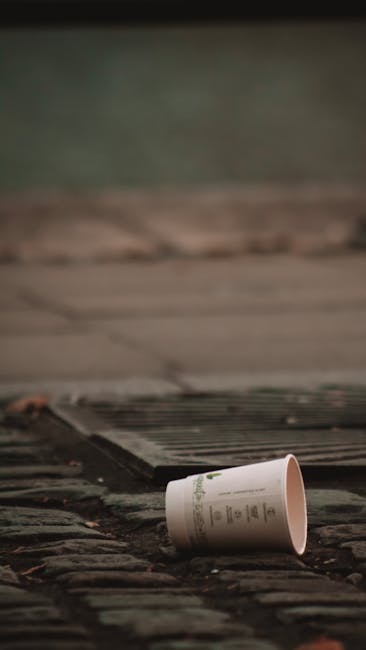 A close-up view of a pile of crushed and crumpled aluminium and plastic soft drink cans, primarily in silver, with some featuring red, blue, and black branding and labels. The cans exhibit various tex